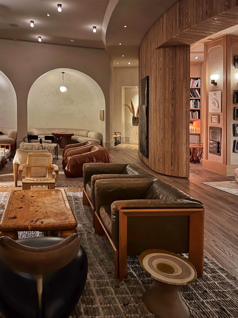 Lobby lounge at Proper Hotel Santa Monica with leather sofas, sculptural lounge chairs, patterned rugs, and a curved wood-paneled wall. Arched seating alcoves, warm lighting, built-in bookshelves, textured walls, and layered wood tables create a collected, residential-style hotel lobby.