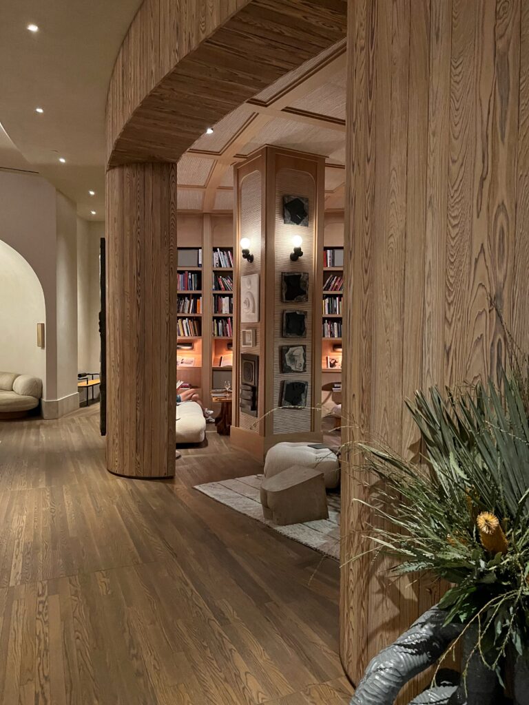 Lobby at Proper Hotel Santa Monica with curved wood-paneled walls, wood flooring, built-in bookshelves, and softly lit lounge seating. A sculptural column, wall-mounted art, rounded furniture, and a large potted plant create a warm, layered hotel interior.