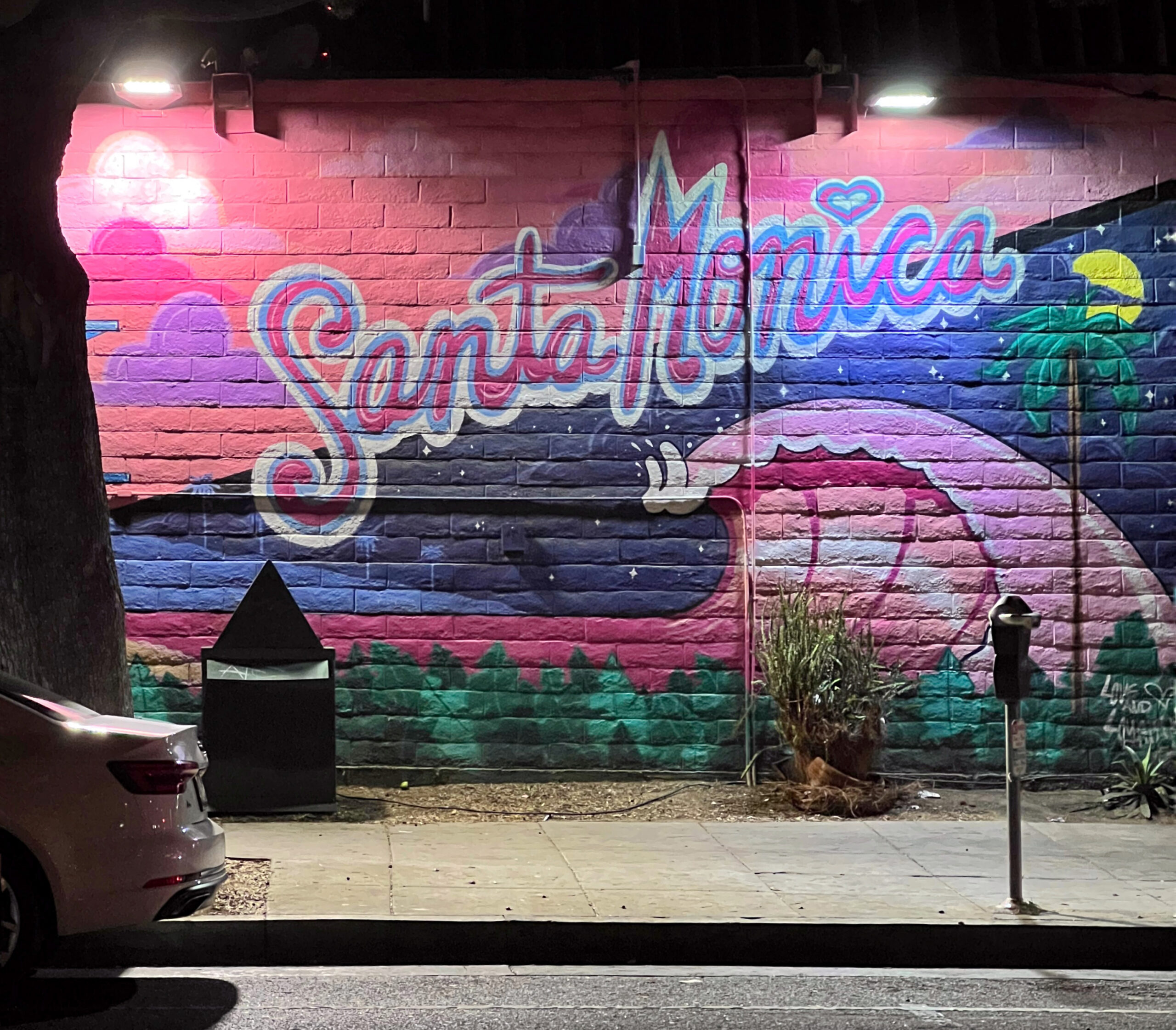 Colorful Santa Monica mural painted on a brick wall at night, with pink, purple, blue, and green landscape details lit by exterior wall lights. A parked car, sidewalk, street, tree, and parking meter frame the urban street scene for an aesthetic Santa Monica travel guide.