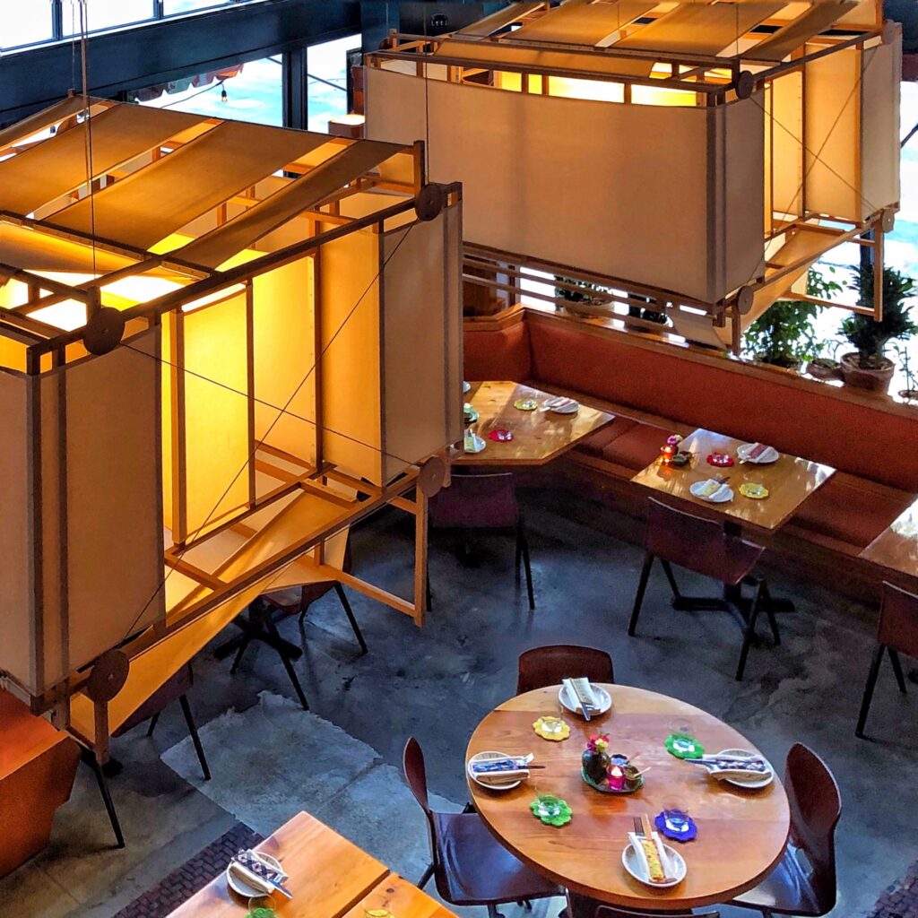 Interior of The Exchange restaurant in Los Angeles, photographed from above with warm suspended light structures, wood tables, orange banquette seating, and colorful table settings. The dining room features polished concrete floors, large windows, potted plants, and softly glowing overhead lantern fixtures.