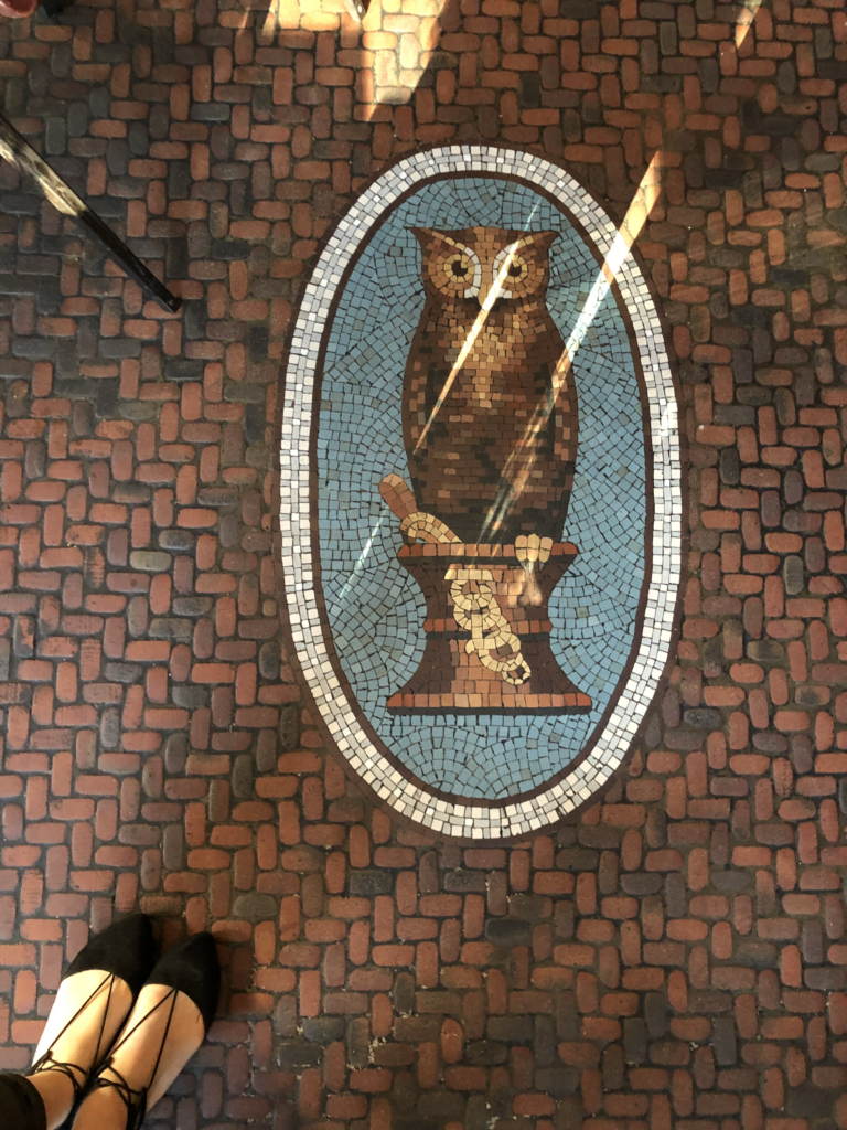 Original drugstore owl mosaic tile detail set into the brick floor at The Exchange restaurant in Los Angeles, photographed from above. The image shows the oval blue-and-brown owl design, surrounding brick tile, angled sunlight, and black lace-up shoes.