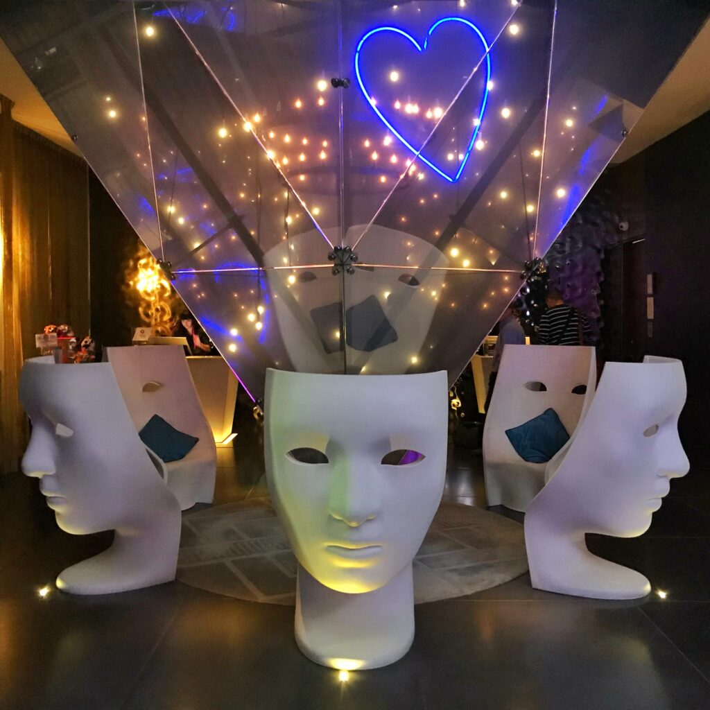 Lobby at W Mexico City with sculptural white face chairs arranged around a faceted glass centerpiece and blue neon heart light. Warm string lights, dark walls, colorful accent pillows, and dramatic uplighting create a playful, modern hotel interior.