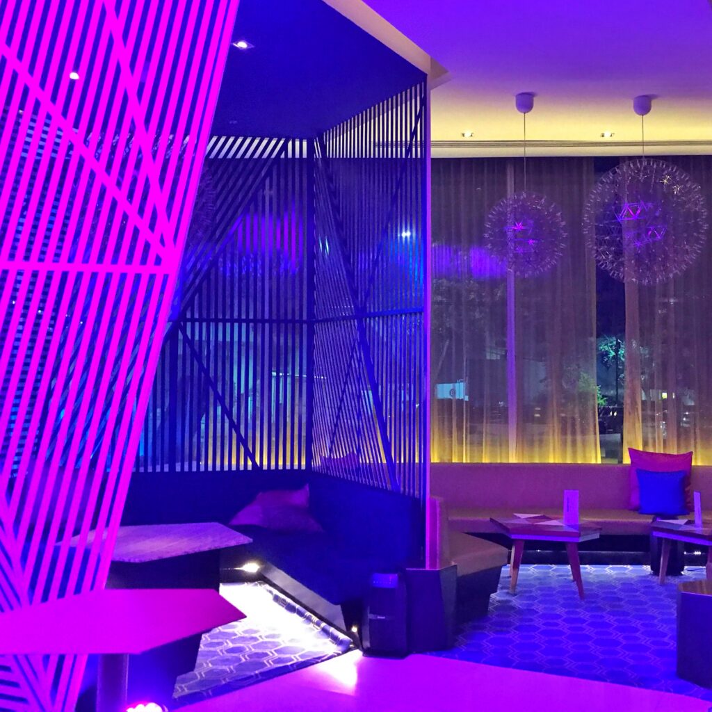 Lobby lounge at W Mexico City with vivid purple and blue lighting, geometric screen partitions, lounge seating, and patterned carpet. Sheer window drapery, sculptural pendant lights, and modern furniture create a colorful nighttime hotel interior.